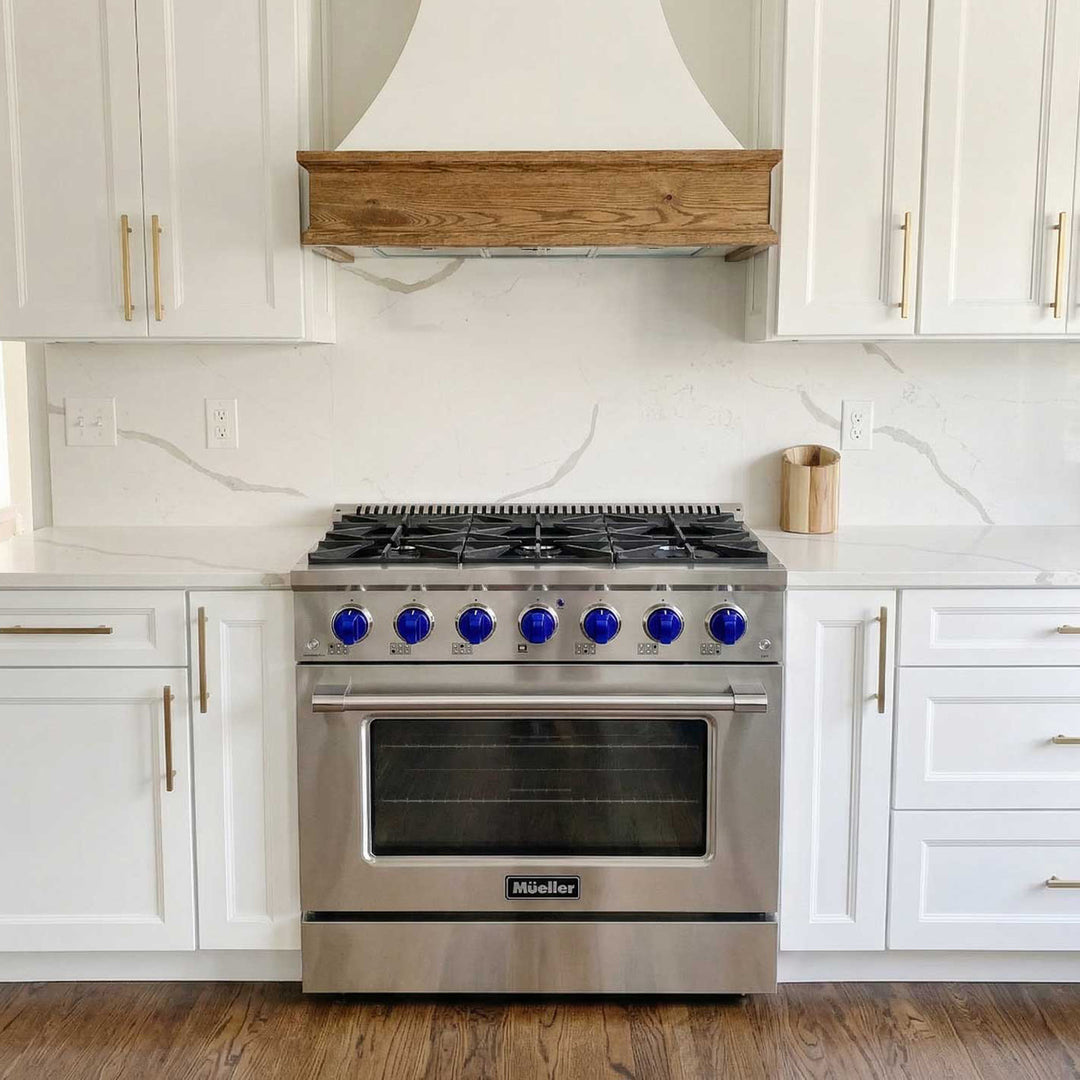 Mueller branded 36 inch stainless steel gas range, with blue knobs, in a white modern kitchen with white cabinets and tiles and a feature range hood.