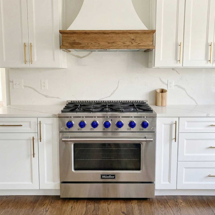 Mueller branded 36 inch stainless steel gas range, with blue knobs, in a white modern kitchen with white cabinets and tiles and a feature range hood.