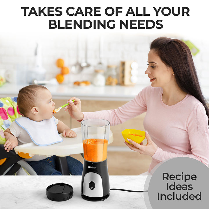Woman feeding a baby with a spoon next to a blender on a kitchen counter.