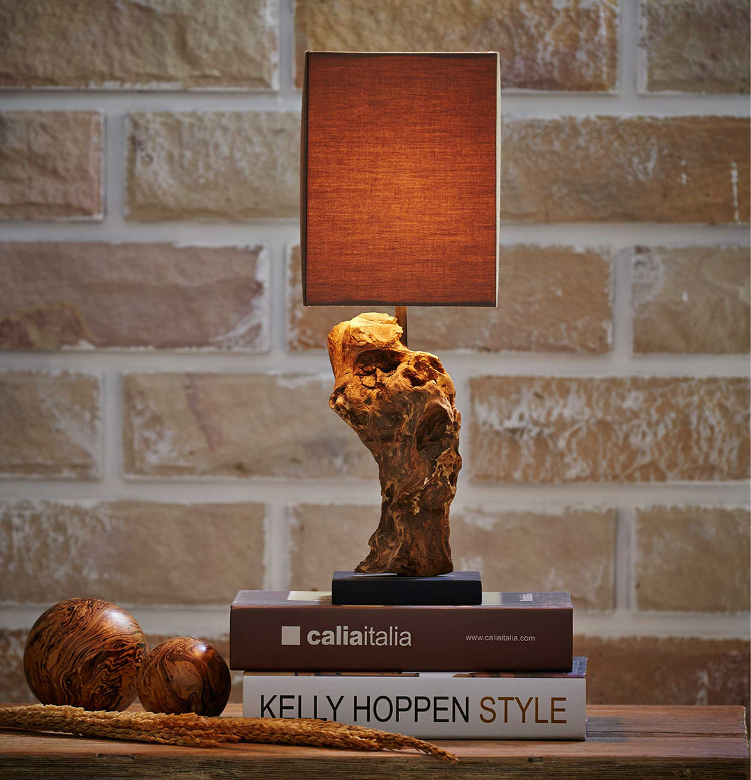 Table lamp with a textured brown shade on top of books against a brick wall.